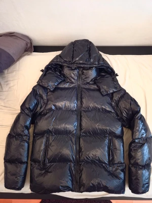 Svart dunjacka - Säljer en svart, glansig dunjacka med stor huva och två dragkedjeförsedda fickor framtill. Jackan har en puffig look och stängs med dragkedja. Perfekt för kalla vinterdagar och riktigt varm tack vare dunfyllningen.