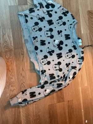 Grå croppad tröja med Mickey Mouse - Säljer en grå croppad sweatshirt med tryck av Mickey Mouse och bokstaven M över hela tröjan. Tröjan har långa ärmar och rund halsringning. Perfekt för dig som gillar Disney och vill ha en avslappnad stil. Den är knappt använd så typ ny och hel