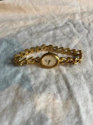 Elegant guldfärgad armbandsklocka - Snygg guldfärgad armbandsklocka med oval urtavla och länkarmband i hjärtformad design. Urtavlan är vit med diskreta markeringar och klockan har en klassisk, feminin look som passar till många stilar. Klockan funkar inte men det går att köpa rätt batterier.