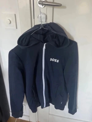 Mörkblå hoodie med dragkedja BOSS - Snygg mörkblå hoodie från BOSS med vit dragkedja och logga på bröstet. Hoodien har huva och två fickor framtill. Perfekt för en avslappnad och stilren look. Det är i barnstorlek!