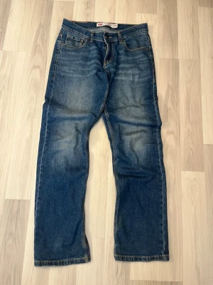 Levi's 511 blå raka jeans - Säljer ett par klassiska blå Levi's 555 jeans med raka ben och fem fickor. Jeansen har snygga slitningar och kontrastsömmar samt den ikoniska läderlappen bak i midjan. Tillverkade i slitstark denim med en tidlös look.