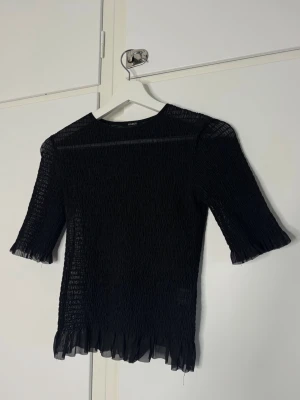 Svart mesh topp med plisserad struktur - Trendig svart mesh topp med plisserad struktur och halvtransparent material. Toppen har trekvartsärm och volangkant nedtill samt vid ärmslut. Perfekt för att lyfta din outfit med en edgy touch.