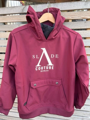Vinröd jacka Slade Couture - Colorado Softshell Snow Hood JR är en riktigt cool och stretchig skidjacka som dessutom är vind- och vattenavisande! Softshell Snow Hood Jr från Slade är en otroligt funktionell snowboardjacka för barn. Med en vattenpelare på 10 000 mm håller den dig torr och skyddad under hela vinteräventyret. Jackan har även en hög andningsförmåga för att du ska kunna hålla dig sval och bekväm. Den justerbara huvan och den höga kragen ger extra skydd mot vinden och kylan.