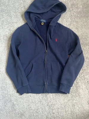 Marinblå hoodie från Polo Ralph Lauren - Snygg marinblå hoodie från Polo Ralph Lauren med dragkedja framtill och klassisk röd logga på bröstet. Hoodien har huva, kängurufickor och ribbade muddar. Tillverkad i mjukt bomullsmaterial som känns skönt mot huden.