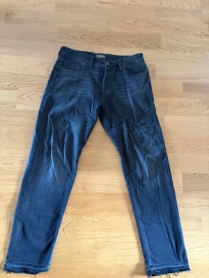 Svarta slitna jeans med råa kanter - Säljer ett par mörkblå jeans med slitningar på både fram- och baksida samt råa, fransiga benslut. Jeansen har klassisk femficksmodell och smal passform. Perfekta för dig som gillar en avslappnad och trendig stil. Storlek: 32/32