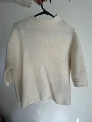 Krämvit stickad tröja med halvpolo - En enkel och stilren krämvit stickad tröja med halvpolo och trekvartsärmar. Tröjan är gjord i ett mjukt, lite luddigt material som känns skönt mot huden. Perfekt att matcha med jeans eller kjol för en clean look.
