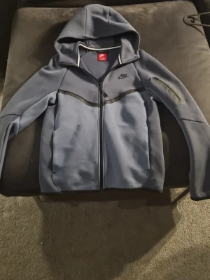 Blå Nike hoodie med dragkedja XS - Snygg blå hoodie från Nike med hel dragkedja och huva. Hoodien har svarta detaljer, två fickor framtill och en extra ficka med dragkedja på ärmen. Perfekt för en sportig och avslappnad stil.