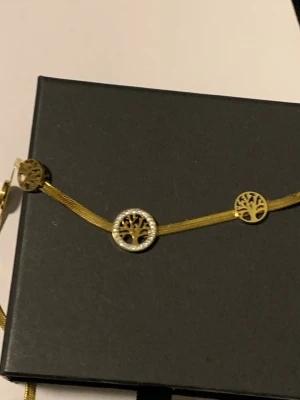Guldigt halsband med livets träd - Snyggt halsband i guldfärgad metall med tre runda berlocker som har motiv av livets träd. Den mittersta berlocken är dekorerad med små gnistrande stenar runt kanten. Perfekt accessoar för dig som gillar detaljer och symbolik.