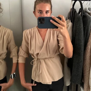Beige omlottblus med knytband - Snygg beige omlottblus med trekvartsärmar och knytband i midjan. Blusen har en djup v-ringning och lätt puffade ärmar för en trendig look. Tillverkad i ett tunt, mjukt tyg som ger ett avslappnat men stilrent intryck.