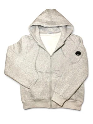 C.P. Company zip hoodie - Säljer en ljusgrå hoodie med dragkedja och huva. Tröjan har ribbade muddar, fickor framtill och en svart patch på ena ärmen. Tillverkad i mjukt sweatshirtmaterial, perfekt för chill dagar. Dragsko i huvan för justerbar passform.