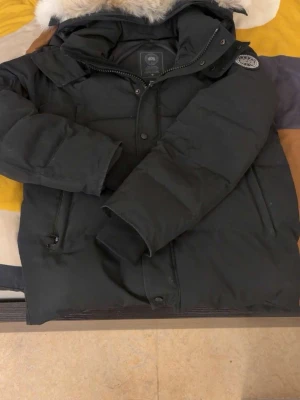 Svart Canada goose vinterjacka med fin päls  - Snygg svart dunjacka med pälskantad huva och patch på ärmen. Jackan har dragkedja och knappar framtill samt två sidofickor med dragkedja. Perfekt för kalla dagar och riktigt varm. Klassisk modell med en modern touch. Storlek s men passar m också