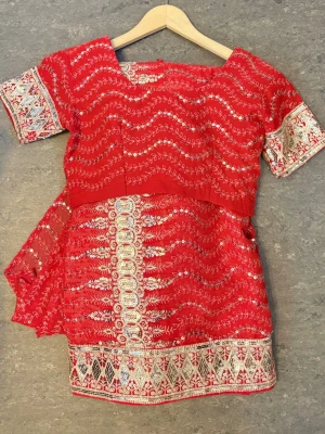 Rött broderat set med blus och kjol (indiska tradition klänning)  - Säljer ett traditionellt rött set med matchande blus och kjol, dekorerat med detaljerat silverfärgat broderi och paljetter. Setet har kortärmad blus med fyrkantig ringning och kjol med bred broderad kant. Materialet är tunt och glansigt, troligen polyester eller liknande.