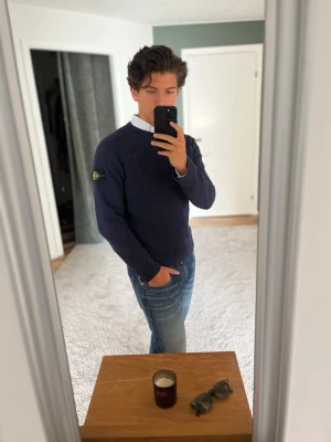 Mörkblå tröja från Stone Island - Säljer en mörkblå rundhalsad tröja från Stone Island med den klassiska logotypen på vänster ärm. Tröjan har ribbade muddar vid ärmslut och nederkant samt en stilren, enkel design. Perfekt att matcha med skjorta under för en clean look. Nopprig men inget som inte går att ta bort met en noppermaskin