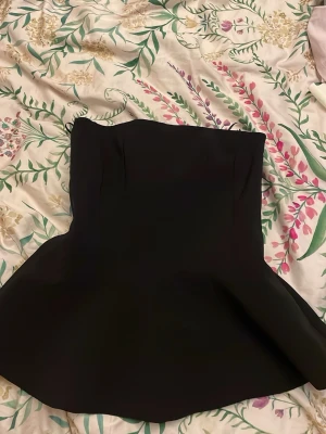 Svart peplum topp från Zara - Säljer en svart peplum topp från Zara i storlek XS. Toppen är axelbandslös med figurnära passform upptill och utställd nertill. Perfekt för att skapa en snygg siluett och passar till både jeans och kjol.