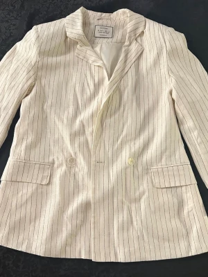 Amisu Vit kavaj med smala svarta ränder M  - Snygg vit kavaj med smala svarta ränder och dubbelknäppning. Klassisk krage och två fickor framtill. Lätt material som passar perfekt till en trendig look. Ärmarna kan bäras både nedfällda och uppkavlade för en mer avslappnad stil.