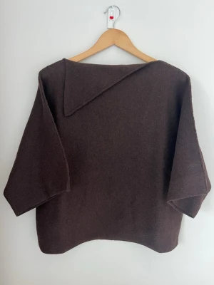Brun stickad kortärmad tröja med vid krage - Mysig brun stickad poncho med breda trekvartsärmar och en unik, asymmetrisk krage. Ponchon har en avslappnad passform och är perfekt för lager på lager. Materialet är mjukt. Använd en gång