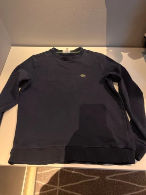 Mörkblå sweatshirt från Lacoste - Mörkblå sweatshirt från Lacoste med klassisk rund halsringning och den ikoniska gröna krokodilloggan på bröstet. Tröjan har ribbade muddar vid ärmslut och nederkant samt en diskret grön detalj på insidan av kragen. Perfekt för en avslappnad stil.