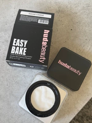 Huda Beauty Easy Bake Sugar Cookie - Loose baking & setting powder från Huda Beauty i nyansen Sugar Cookie. Kommer i en fyrkantig burk med svart lock och rosa logga. Pudret är ljust och ger en matt finish, perfekt för att fixera basen och highlighta ansiktet.
