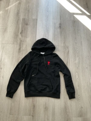 Svart hoodie med rött hjärta A - Snygg svart hoodie med huva och känguruficka. På bröstet sitter en röd broderad detalj med ett hjärta och bokstaven A. Hoodien har ribbade muddar vid ärmslut och nederkant samt dragsko i huvan. Perfekt för en chill och stilren look.