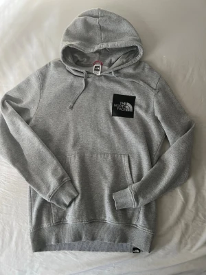 Grå hoodie från The North Face - Snygg grå hoodie från The North Face med klassisk logga på bröstet. Tröjan har huva med dragsko, magficka och ribbade muddar vid ärmar och nederkant. Tillverkad i mjukt bomullsmaterial som är skönt att ha på sig.