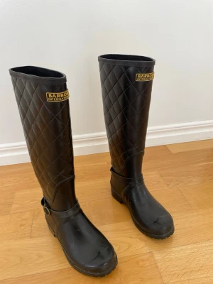 Svarta höga stövlar från Barbour - Snygga svarta höga stövlar från Barbour International med quiltat mönster och spänndetalj vid ankeln. Tillverkade i gummi, perfekta för regniga dagar. Klassisk och stilren design med logga framtill.