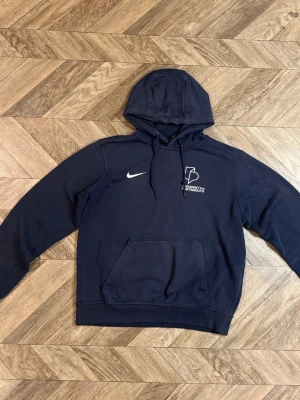 Mörkblå Nike hoodie - Mörkblå hoodie från Nike med huva och magficka. Tryck på bröstet med texten 'University of Portsmouth' och en vit Nike-logga. Klassisk passform och ribbade muddar. Perfekt för en avslappnad och sportig stil.