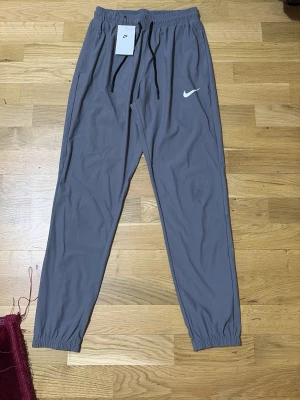 Grå Nike mjukisbyxor med dragsko - Snygga grå mjukisbyxor från Nike med svart dragsko i midjan och elastiska benslut. Byxorna har en klassisk Nike-logga på benet och är tillverkade i ett mjukt och lätt material som känns skönt mot huden. Perfekta för chill eller träning.