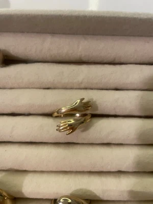 Guldfärgad ring med handmotiv - Unik guldfärgad ring i metall med två händer som möts framtill. Ringen har en öppen design och glänsande finish. Perfekt för dig som gillar kreativa och konstnärliga accessoarer.