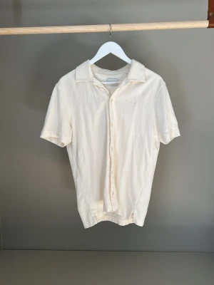 Krämvit kortärmad skjorta från Gant i frottéliknande material - Stilren krämvit skjorta med korta ärmar och klassisk krage. Skjortan har knäppning framtill och är tillverkad i ett mjukt material som känns skönt mot huden. Perfekt för en avslappnad och fräsch look.