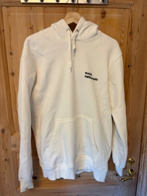 Vit hoodie från Mads Nørgaard - Säljer en stilren vit hoodie från Mads Nørgaard med broderad logga på bröstet. Hoodien har stor huva med dragsko, magficka och ribbade muddar vid ärmslut och nederkant. Perfekt för en avslappnad och trendig look.