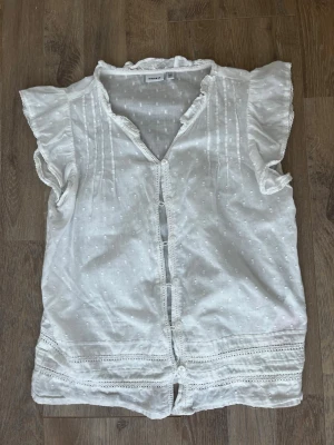 Vit blus med volangärm från Name It - Söt vit blus från Name It med korta volangärmar och knappar framtill. Blusen har diskret prickstruktur, dekorativa spetsdetaljer och rynkade partier vid axlarna. Perfekt till sommaren och lätt att matcha med jeans eller kjol💗