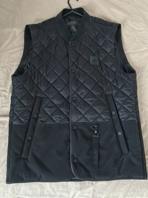 Svart quiltad dunväst från Moose Knuckles - Snygg svart dunväst från Moose Knuckles med quiltat mönster och tryckknappar framtill. Västen har två fickor med knappstängning och en diskret logga på bröstet. Perfekt lager på lager-plagg för kyliga dagar.