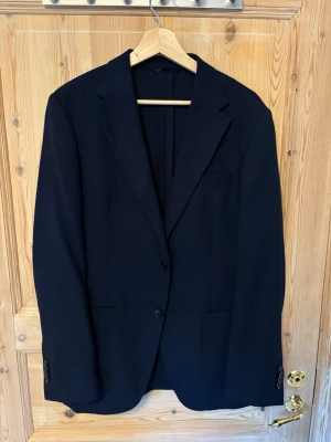 Klassisk svart kavaj från Volt - Stilren svart kavaj med två knappar framtill och klassiska slag. Kavajen har två framfickor och en bröstficka. Passar perfekt till skjorta och jeans för en snygg look. Materialet är en mönstrad textil, troligtvis ullblandning.