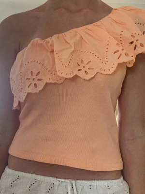 Persikofärgad oneshoulder topp Zara - Säljer en persikofärgad oneshoulder topp från Zara med bred volang och broderade blommor längs kanten. Toppen är ribbad och har en snygg, croppad passform som passar perfekt till sommaren.