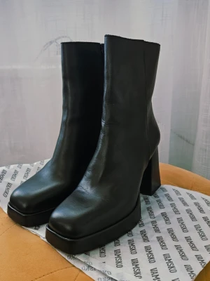 Svarta platåstövletter från Vamsko st 38 - Snygga svarta stövletter från Vamsko med chunky platå och bred klack. Tillverkade i slätt skinn med fyrkantig tå och dragkedja på insidan. Perfekta för dig som vill ha en edgy och trendig look. Ny pris 2800 kr. Köparen står för frakt. Ej retur. 