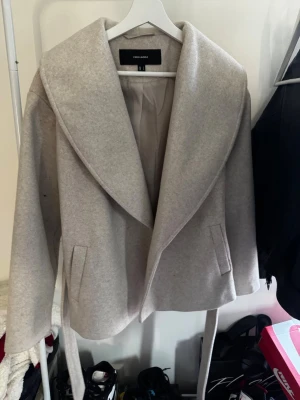 Beige rock med bälte från Vero Moda - Snygg beige rock från Vero Moda med bred krage och bälte i midjan. Rocken har en klassisk, stilren look och är perfekt för kyligare dagar. Långa ärmar och två fickor framtill ger både stil och funktion.