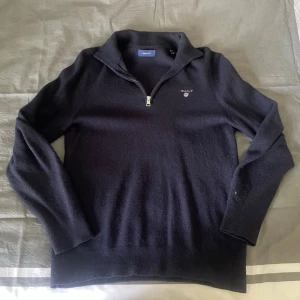 Svart halvzip-tröja från GANT - Klassisk svart stickad tröja från GANT med halv dragkedja och broderad logga på bröstet. Tröjan har hög krage och långa ärmar, perfekt för lager på lager. Materialet känns mjukt och bekvämt, och passformen är avslappnad men ändå stilren.⁉️Skriv vid frågor⁉️
