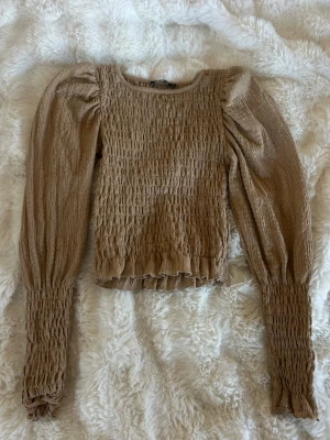 Beige smockad topp med puffärm - Trendig beige topp med smockad struktur och långa puffärmar. Modellen är croppad med volangkant nedtill och vid ärmslut. Perfekt för dig som gillar en feminin och cool stil. Materialet känns lätt och stretchigt.