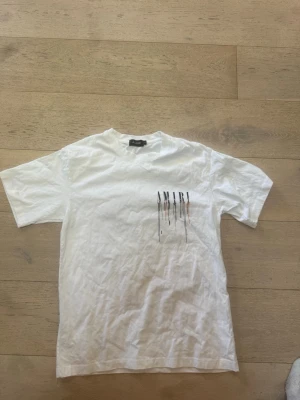Vit AMIRI t-shirt med tryck - Vit t-shirt från AMIRI med färgstarkt tryck på bröstet och ryggen där bokstäverna har droppande effekt i flera färger. Klassisk passform och rund halsringning. Perfekt för dig som gillar streetwear och vill sticka ut.