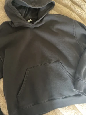 Svart basic hoodie med huva - En enkel och stilren svart hoodie med huva och stor magficka framtill. Hoodien har ribbade muddar vid ärmslut och nederkant. Perfekt för chill dagar eller när du vill ha en avslappnad look.