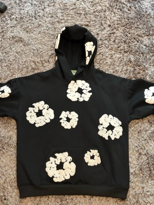 Svart hoodie med vita blommor Denim Tears - Säljer en svart hoodie från Denim Tears med stora vita blomliknande tryck över hela tröjan och huvan. Hoodien har magficka och ribbade muddar. Perfekt för dig som vill sticka ut med en unik och trendig stil.