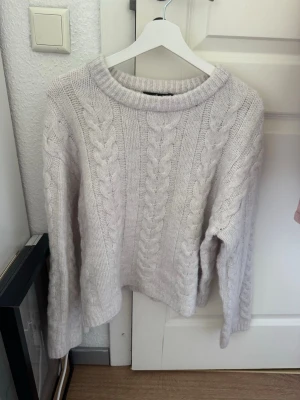 Ljusgrå kabelstickad tröja - Mysig ljusgrå kabelstickad tröja med rund halsringning och långa ärmar. Tröjan har ett klassiskt flätmönster framtill och är tillverkad i ett mjukt, tjockt material som passar perfekt för kyligare dagar.