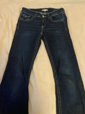 Mörkblå bootcut jeans med vita sömmar - Säljer ett par mörkblå bootcut jeans med tydliga vita kontrastsömmar. Jeansen har fem fickor, knappgylf och dekorativa ficklock bak med knappar. Klassisk denimlook som passar till det mesta.det är som en svag linje längst ner eftersom de har varit uppsydda men inget man tänker på. I storlek 164