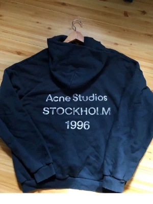 Svart hoodie Acne Studios Stockholm - Svart hoodie från Acne Studios med tryck på ryggen där det står 'Acne Studios STOCKHOLM 1996'. Klassisk passform med huva och ribbade muddar. Perfekt för en avslappnad och trendig streetwear-look.