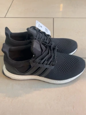 Svarta Adidas Ultra Boost  - Snygga svarta Adidas Ultra Boost sneakers med stickad ovandel i mesh för extra andningsförmåga. Skorna har klassiska tre ränder på sidan, svart snörning och en vit Boost-sula som ger grym komfort. Perfekta för dig som gillar stilrena och sportiga sneakers.