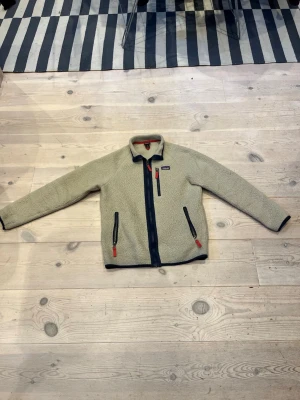 Patagonia fleece 13-14yr  - Mysig beige teddyjacka med svarta dragkedjor och röda zippar. Jackan har hög krage, tre fickor framtill och kontrastfärgade detaljer längs kanterna. Perfekt för kalla dagar och ger en avslappnad, trendig look.