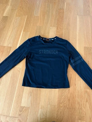 Svart långärmad träningströja Stronger - Snygg svart långärmad träningströja från Stronger med tryckt logga på bröstet och diskreta ränder på ena ärmen. Tillverkad i ett stretchigt funktionsmaterial som passar perfekt till träning och sportaktiviteter. Nästan aldrig använd 