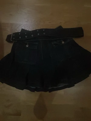 Svart minikjol med fickor och bälte - Säljer en svart minikjol i jeansmaterial med två stora fickor framtill och ett brett bälte med metallhål. Kjolen har en lätt utställd, plisserad form som ger en cool och edgy vibe. Perfekt för dig som gillar streetstyle och vill sticka ut.
