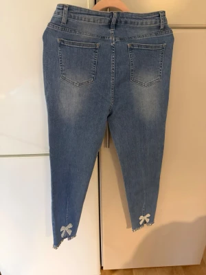 Blå jeans med pärlrosetter - Säljer ett par ljusblå jeans med raka ben och två bakfickor. Nedtill på benen finns dekorativa rosetter av pärlor och strass, samt pärl- och strassdetaljer längs fållen. Jeansen har hög midja och är i klassisk denim utan synligt märke. Jätte mycket stretch- mjuka och sköna. Lite kortare modell. 