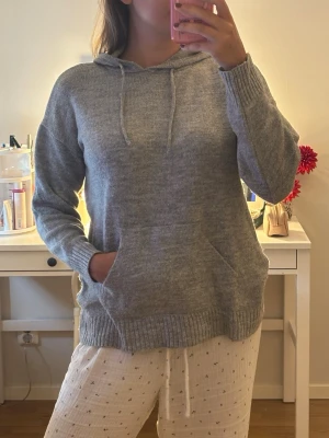 Grå stickad hoodie med ficka - Mysig grå stickad hoodie med huva och dragsnören. Tröjan har en magficka och ribbade muddar vid ärmslut och nederkant. Perfekt för chill dagar eller när du vill ha något bekvämt och stilrent.